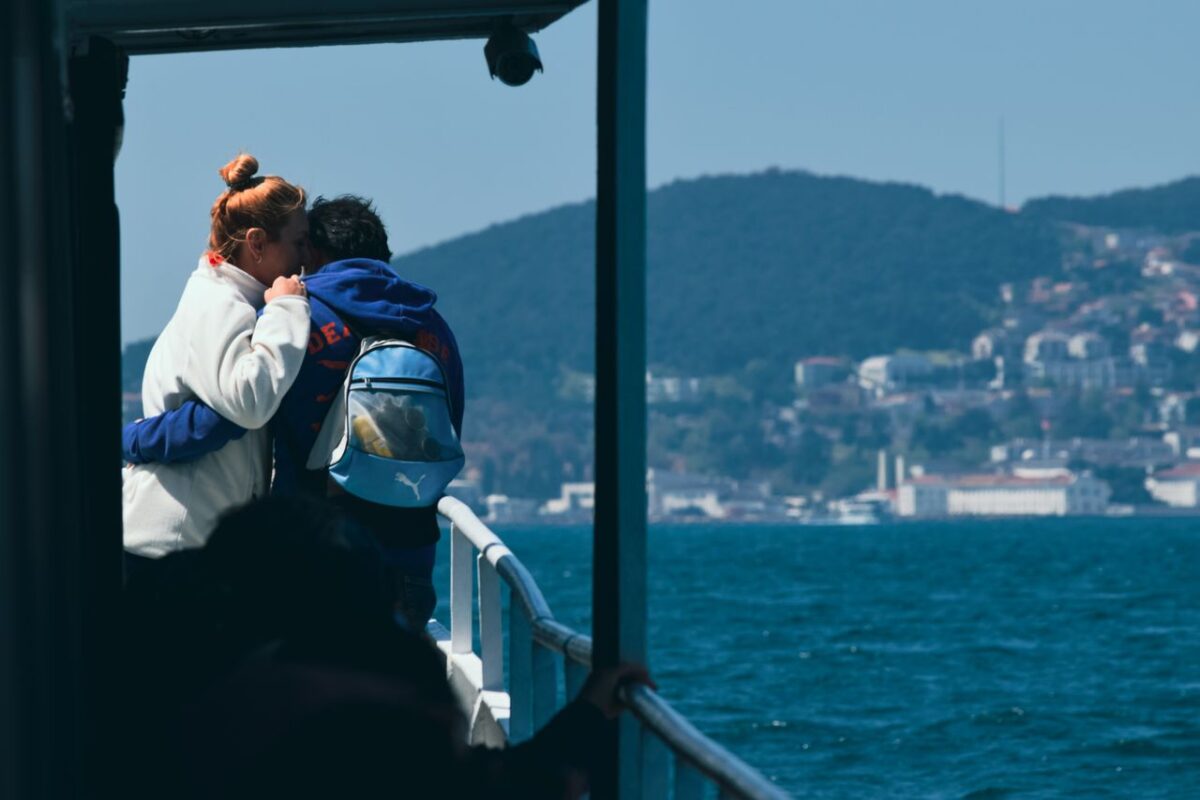 A couple embraces on a ferry, overlooking the ocean with a scenic coastal backdrop.