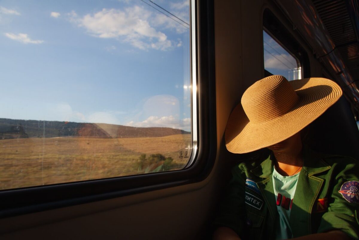 A person wearing a straw hat rests by a train window, enjoying the scenic countryside views.